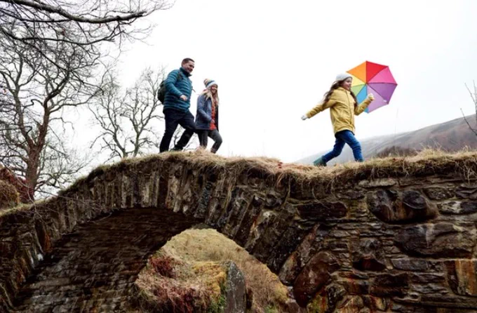 A family group outdoors compromising of two adults and a child in winter clothing walking over a bridge 