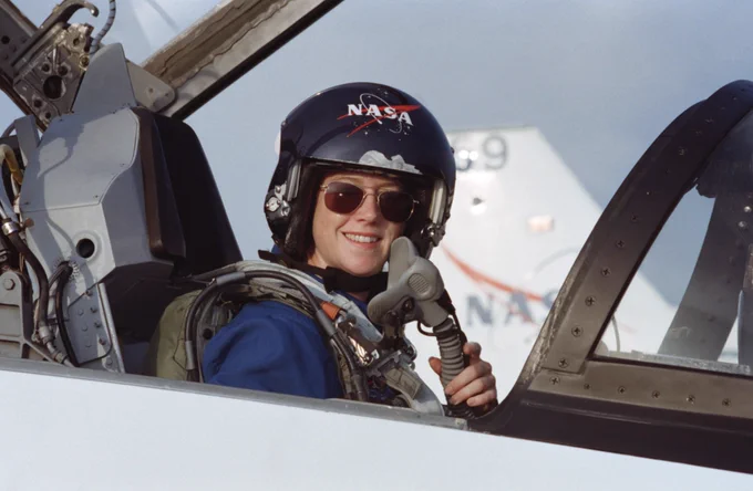 Pam Melroy, seated in the forward station of a T-38 jet trainer in 2000 prior to joining other astronauts to train for a mission to the International Space Station. She is wearing sunglasses and a helmet and is smiling at the camera. Credit: NASA