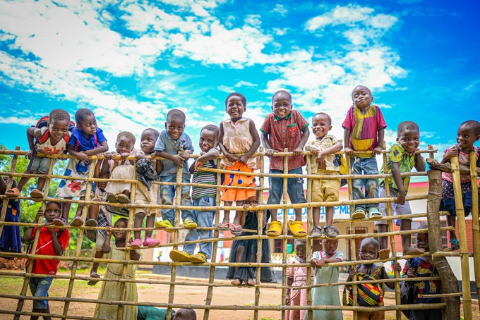 A group of children smiling and playing on a colorful playground structure under a clear blue sky.