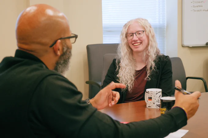 A young woman with albinism smiles as a man speaks to her with a pen in his hand at an office conference table, which has two coffee mugs and a piece of paper on it.