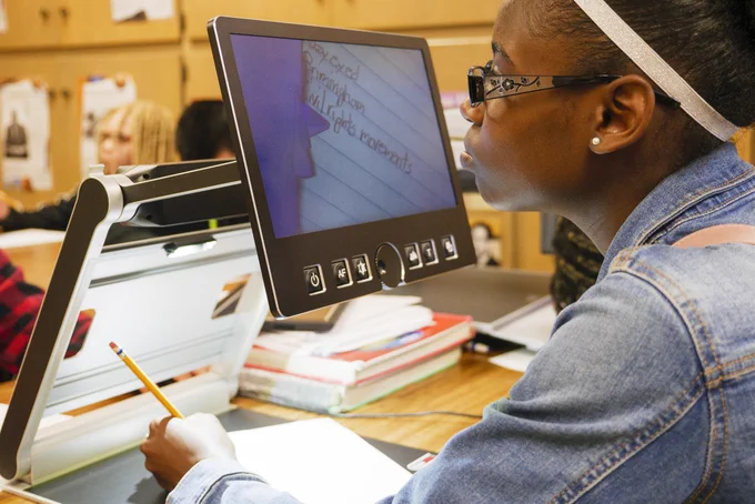 A young woman in a classroom listens intently as she uses a large magnifier to take notes on the Civil Rights Movement.