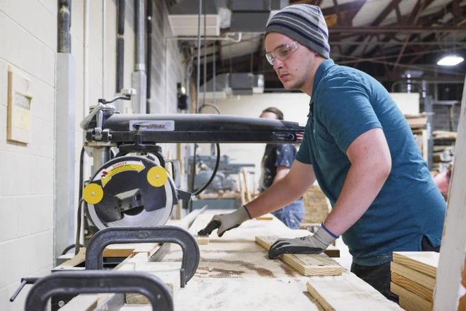 A young man wearing protective eyewear and gloves prepares to use a woodworking saw in a workshop.
