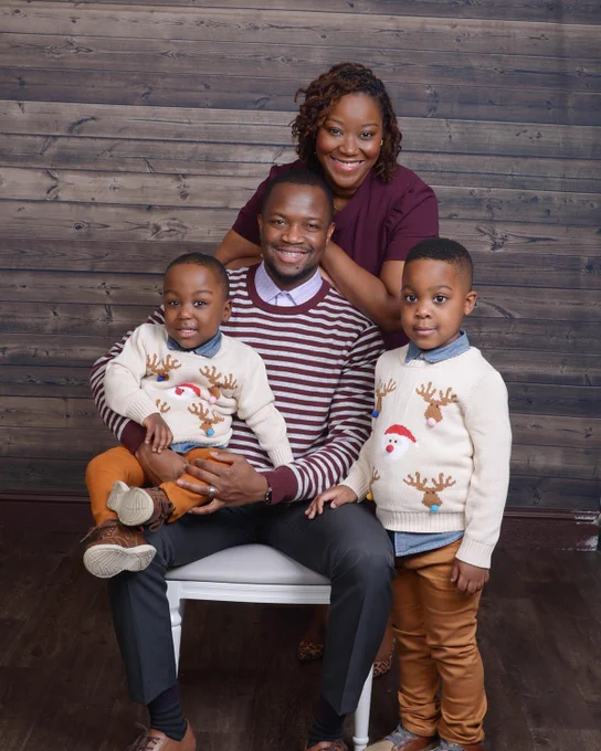 Family portrait featuring four smiling individuals in coordinated outfits, two adults and two children, posing in a studio with a wooden backdrop.