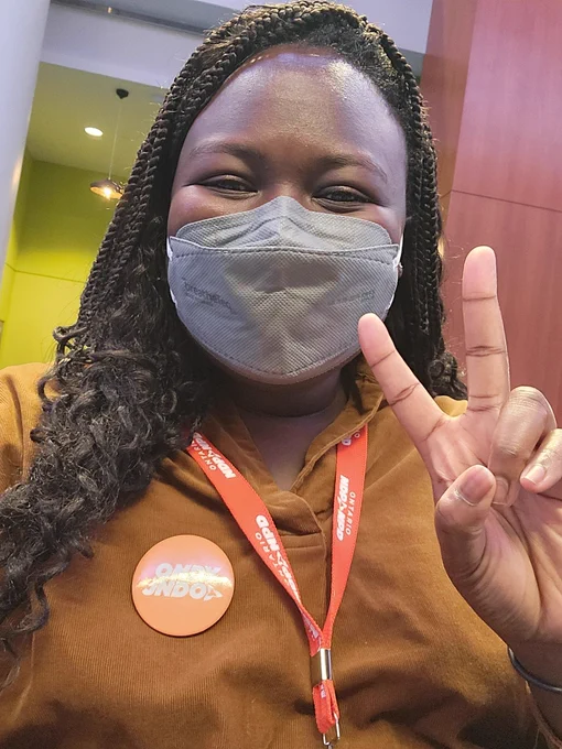 Akuah is wearing a grey mask and a brown collard shirt with an orange ONDP logo lanyard and an orange ONDY/JNDO button with white text. She is holding up a peace sign. 