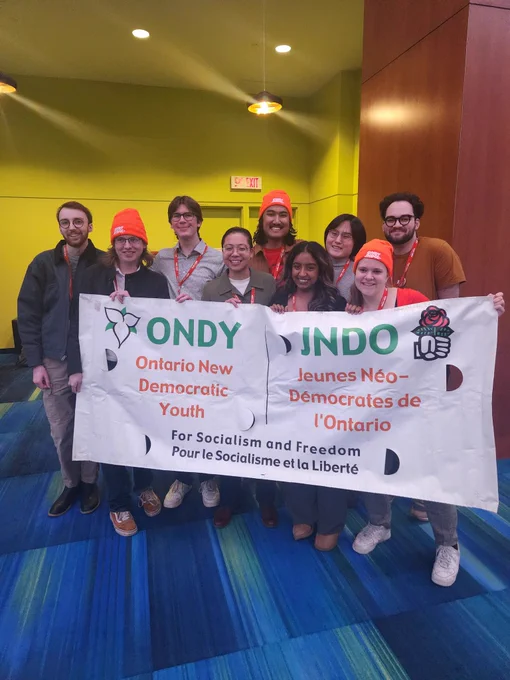 A number of youth are holding up a white banner with the ONDY/JNDO logo in green and orange text and the slogan in black text. There is a picture of a trillium on the top left and a white hand holding a rose on the top right. 