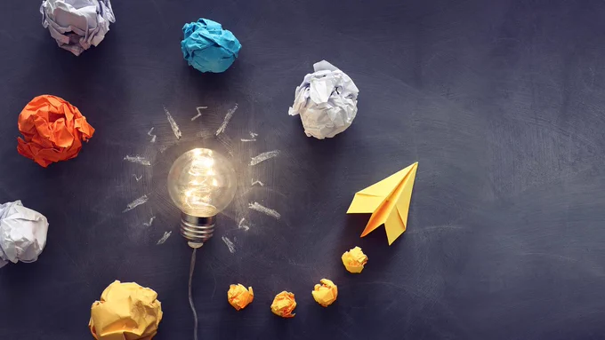 A lightbulb on chalkboard surrounded by crumpled paper leading up to a paper airplane.