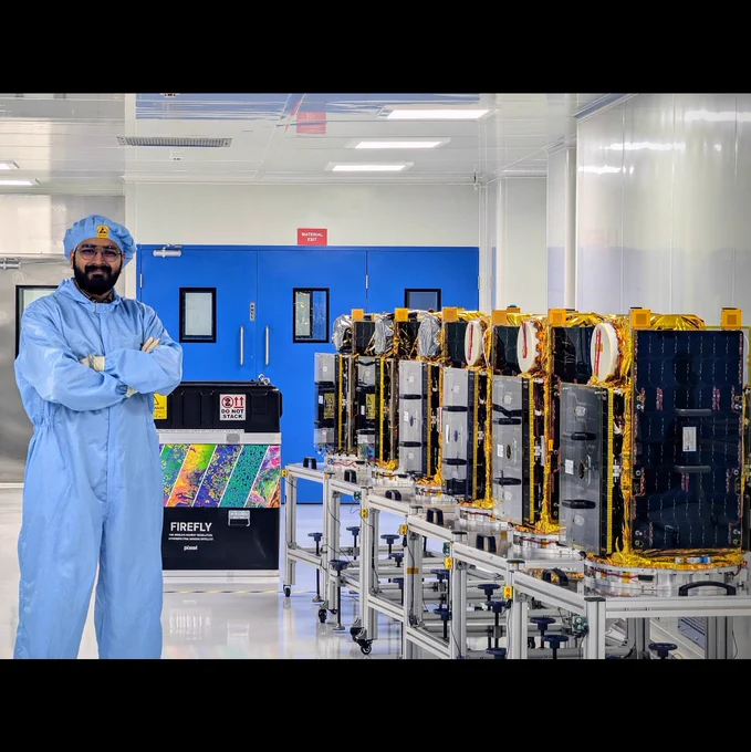Author in clean-room atitre, standing besides six shiny satellites, with a colorful container lid in the background. 
