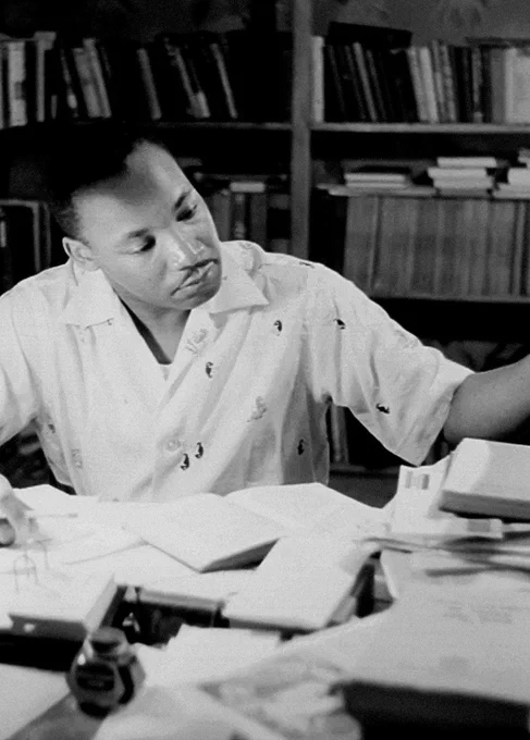 A black and white photo of Dr. Martin Luther King Jr. sitting at a desk, looking down at paperwork and books spread out in front of him. Behind him is a bookshelf full of books.