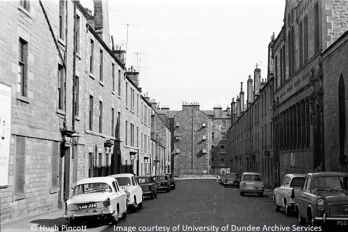 Black and white photograph looking up a street. c 1960s cars are parked on both sides. 3 storey tenements with gas lamps at the doors are on the left  and further back on the right. Nearer on the right is a building with engraved lettering indicating it is Balfour Street Public School. At the end of the street is tenement rear.
