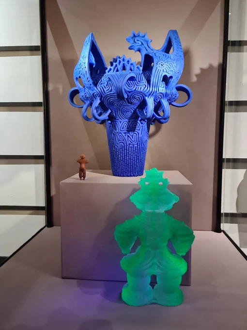 A display of Jomon objects, 'JOMON Vitrified' DUROM.2022.364 A green glass sculpture. 'Dogu Capsule Toy' a small brown plastic figure. 'Jomon (Blue)' DUROM.2017.332 A large bright blue vase, decorated with lines, handles and markings.