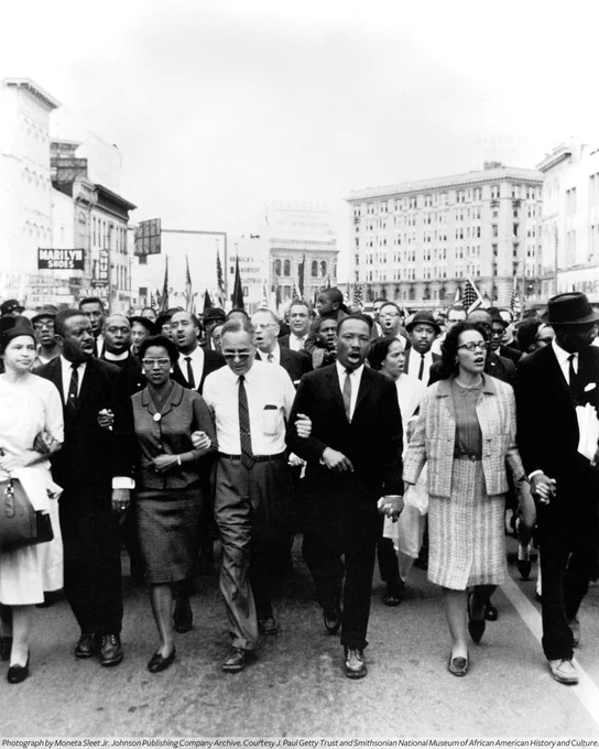 A black-and-white photograph of Dr. Martin Luther King leading a group of protestors during the Selma to Montgomery March in 1965 alongside his wife Coretta and fellow activists Rosa Parks, Ralph Abernathy, his wife Juanita, Dr. Ralph Bunche, and Fred Shuttlesworth.