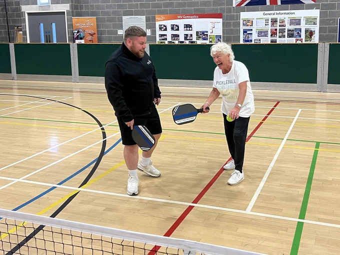 Two individuals playing pickleball indoors, standing on either side of a net with paddles and a ball visible.