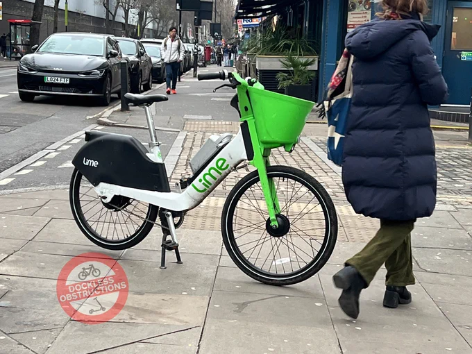A pedestrian navigates around a Lime Bikes abandoned broadside on a pavement in front of a pedestrian crossing in Camden.