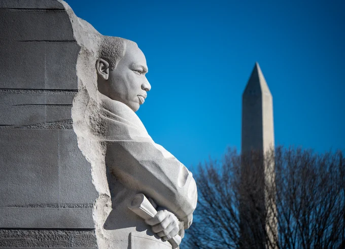 The Martin Luther King, Jr. Memorial in Washington. The Stone of Hope, a granite statue of King, is seen from the side; King's head and upper body, as seen in profile, appear to emerge from the rock. The sky behind the statue is completely clear; a barren tree and the Washington Monument, both out of focus, stand out in the background. Credit: NASA/Bill Ingalls