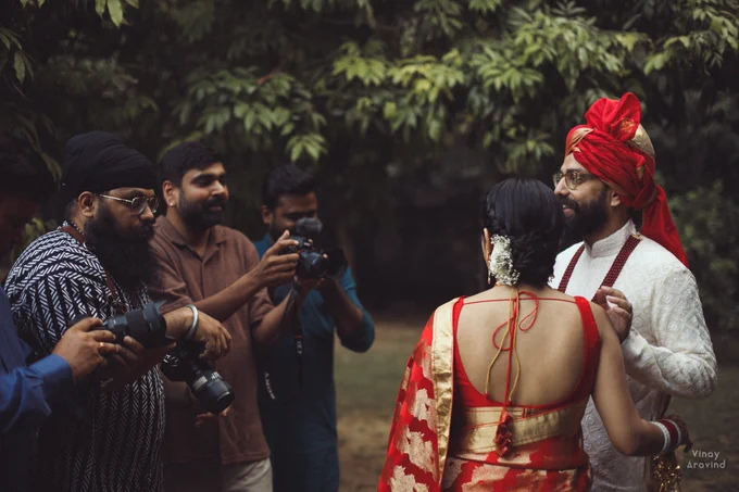 A green outdoor setting with a group of photographers to the left of the frame, shooting a couple in red and white, on the right of the frame. The groom is laughing. 