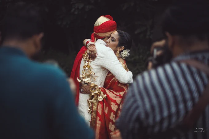 A green outdoor setting with a couple in red and white hugging each other. In the foreground are the backs of two photographers, taking the same shot, out of focus. 