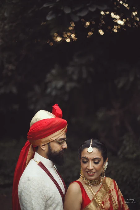 A green outdoor setting with a head-and-shoulders shot of the couple in red and white, where they occupy the lower part of the frame. The bride's face is towards the camera, but she's looking down. The groom, on the left is looking at the bride. Behind them and above them in the frame is foliage, with some out-of-focus highlights. 