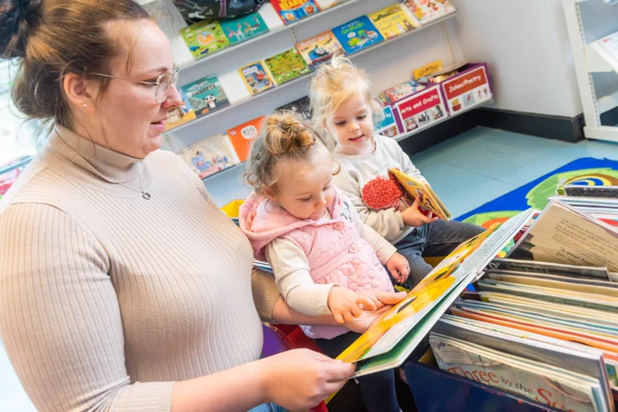 A lady is kneeling next to 2 small children showing them a picture book in a library 