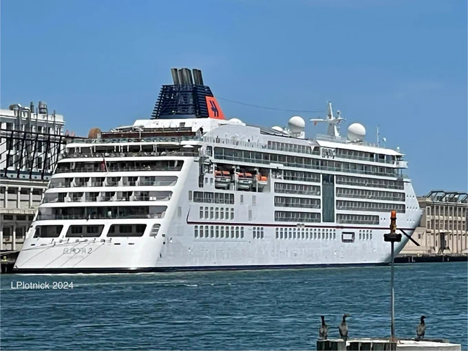 The Europa 2 luxury cruise ship in port with her aft and starboard sides in view. She is a smallish ship, all white with a dark blue funnel and has what appears to be 7 decks featuring private balconies and large panoramic windows. The ship’s stern is tiered with open deck spaces, and lifeboats are visible along the side.