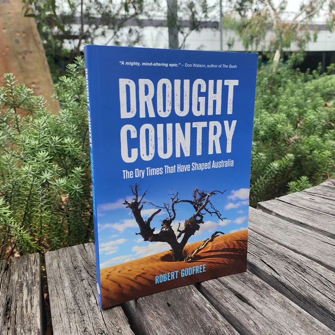 The book Drought Country sitting on a wooden boardwalk surrounded by native vegetation. The book cover depicts a dead tree in a sandy desert under blue skies.