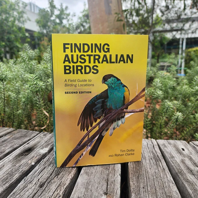 The book Finding Australian Birds, Second Edition, sitting on a wooden boardwalk surrounded by native vegetation. The book cover features a hooded parrot perched on a branch with its wing extended.
