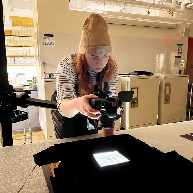 A person adjusting a camera mounted on a tripod, focusing on a small, illuminated object on a tabletop in a well-lit room with filing cabinets
