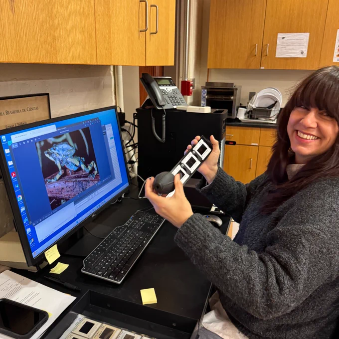 A person holding a slide and using a computer in a laboratory setup, smiling at the camera. The screen displays a colorful microscopic image.