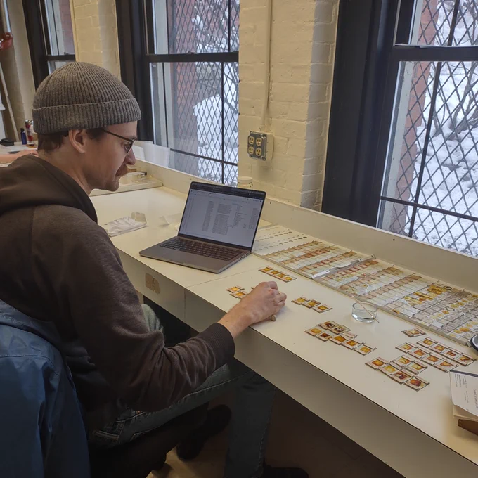 An individual examines microscope slides beside a laptop on a table near a window, in a room with brick walls and a snowy view outside.