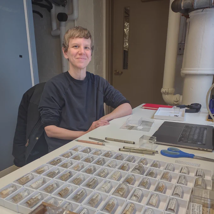 Person smiling while seated at a desk with organized trays of dry stony coral specimens, a laptop, and office supplies, in a laboratory setting.