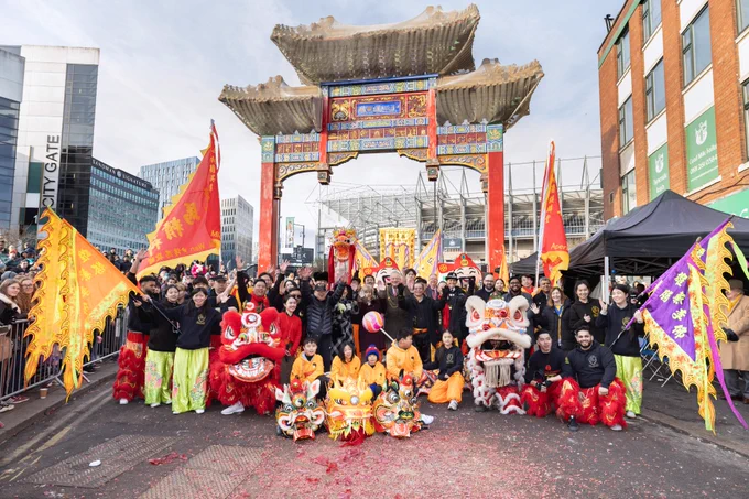 Group photo of some of the performers in traditional, colourful attire, holding flags with Lord Mayor of Newcastle, Cllr Rob Higgins in the middle. Chinese Arch is the backdrop with St James' Park stadium visible behind.