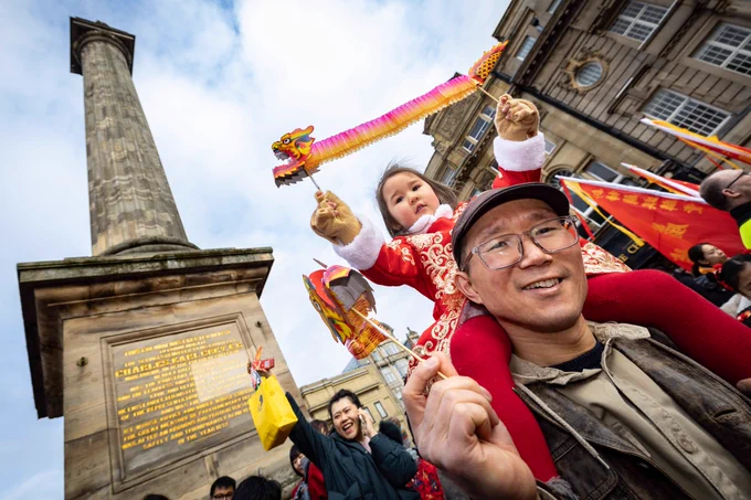 A young child is sat on a man's shoulders and she's holding a paper dragon. Grey's Monument is behind.
