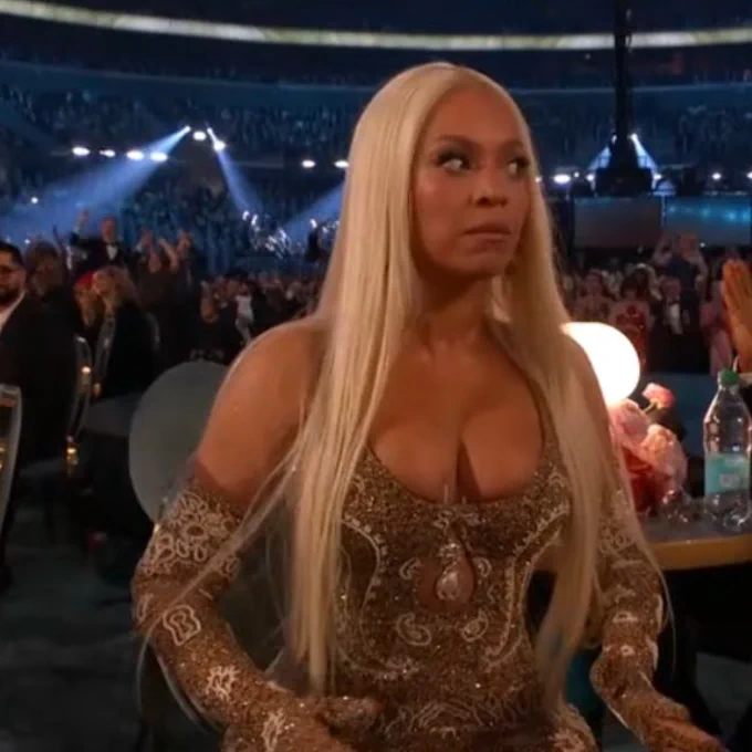 Beyoncé sits at a table at the Grammy Awards, wearing a shimmering gold gown with intricate embellishments and matching gloves. She has long, straight blonde hair and a shocked or surprised expression while looking off to the side.