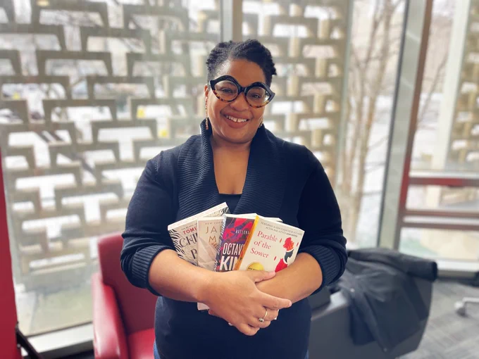 Crystal Kerr with short black hair and glasses wearing a blue sweater stands in Mills Memorial Library holding books written by Black authors.