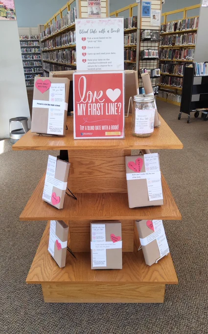 Books covered in brown paper on a display shelf at the Bellefonte Library. The books have hearts denoting genre, the first line of the book, and a book mark attached to them. A sign says: Love my first line? Try a blind date with a book!