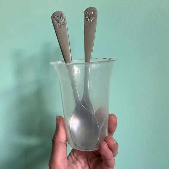 Image of hand holding a plastic cup with two metal spoons in front of a green background