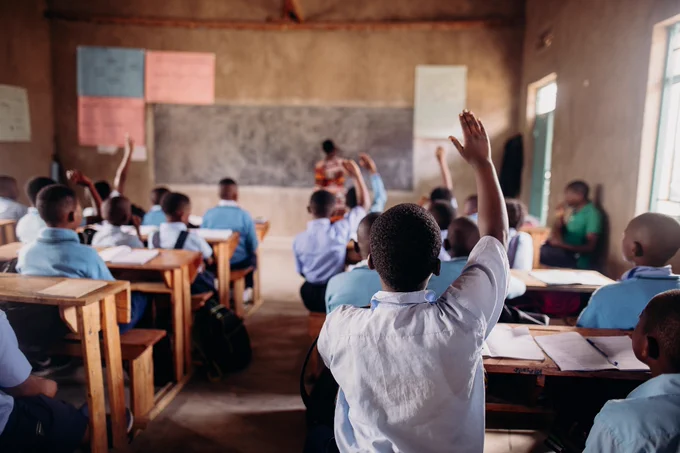 A classroom scene with students in uniform seated at desks, with one student raising a hand. A teacher stands by the chalkboard facing the class.