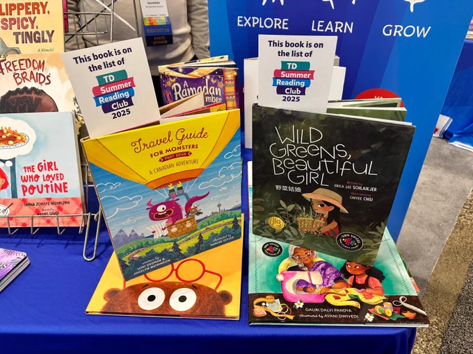 A display of children's books at OLA Super Conference, showcasing titles from Sleeping Bear Press, including two selections from the TD Summer Reading Club 2025 list. These two books are prominently featured on stands: Travel Guide for Monsters: Part Deux - A Canadian Adventure by Lori Degman, illustrated by Marcus Cutler, with a colorful cover depicting a friendly purple monster traveling across Canada; and Wild Greens, Beautiful Girl by Erica Lee Schlaikjer, illustrated by Chinyere Chu, featuring a young girl wearing a straw hat and gathering greens, with a warm, nature-inspired color palette. Signs above the books confirm their inclusion in the 2025 TD Summer Reading Club list. Other books, including The Girl Who Loved Poutine and Freedom Braids, are partially visible in the background. The display is set up on a blue tablecloth with a backdrop featuring the words "EXPLORE," "LEARN," and "GROW."