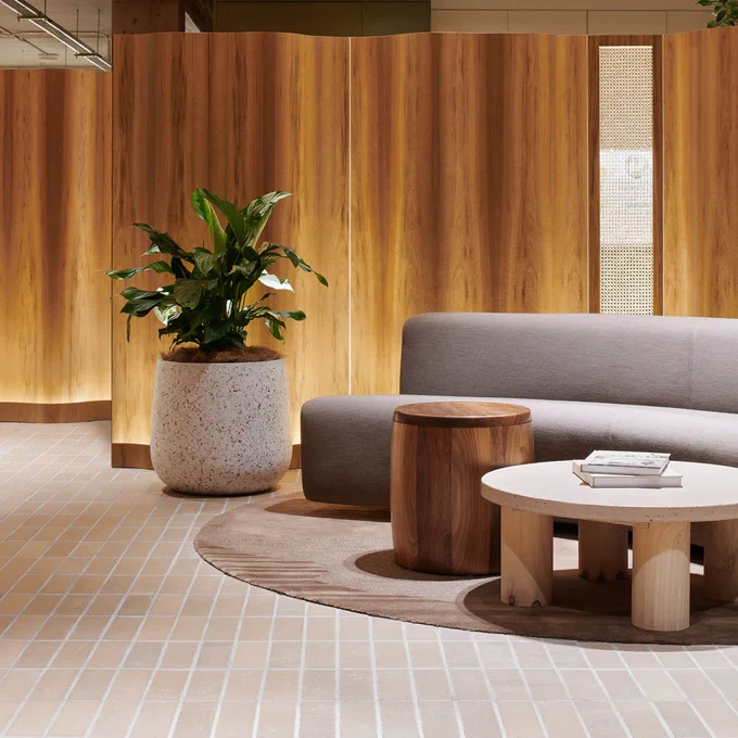 Modern lounge area with a light gray sofa, wooden coffee table, side stool, and a large plant in a white pot. The space features wooden walls and a patterned cream floor.