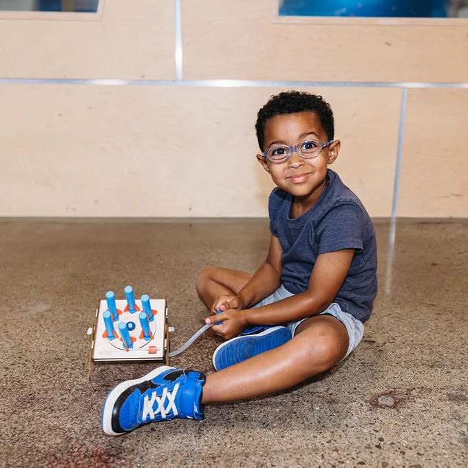 A child wearing glasses sits on the floor, holding a CrunchLabs Rocket Revolver. It is a wooden toy with foam rockets facing the camera.