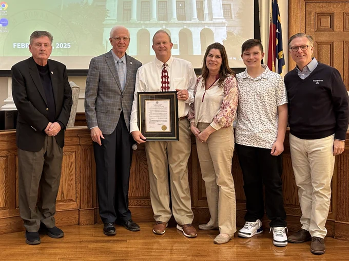 Man holds framed copy of proclamation while posing with his wife, son, and three elected officials