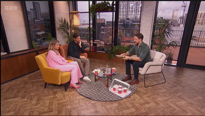 Three people sitting and talking in a TV studio with a cityscape view in the background, on the set of a BBC show.