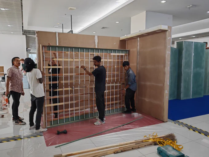 A group of workers assembling a wooden exhibition booth structure indoors. They are handling a wooden panel with a grid framework, possibly for lighting or display purposes. The floor is covered with protective sheets, and various tools, including a drill, broom, and ropes, are scattered nearby. The background features other booth structures in different stages of assembly, with a bright and modern exhibition hall setting.