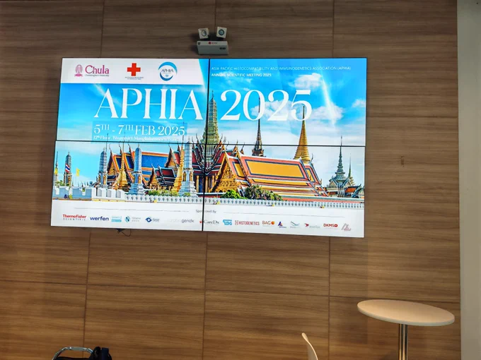 A large digital screen displaying a promotional banner for the APHIA 2025 Annual Scientific Meeting. The banner features an image of a Thai temple with golden roofs and intricate details, set against a bright blue sky. The event details mention it will be held from February 5th to 7th, 2025, at the 12th floor of the Bhumpasit Mongkolsawat Building. Logos of sponsors and organizers, including Chulalongkorn University and the Asia Pacific Histocompatibility and Immunogenetics Association (APHIA), are visible at the top and bottom of the screen.