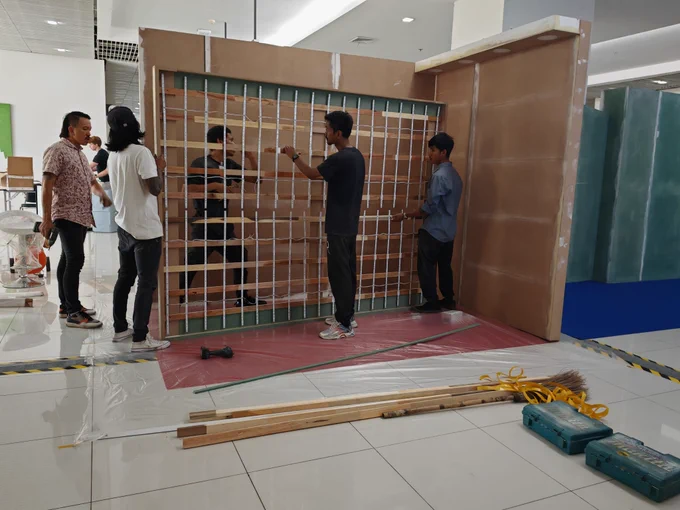 A group of workers assembling a wooden exhibition booth structure indoors. They are handling a wooden panel with a grid framework, possibly for lighting or display purposes. The floor is covered with protective sheets, and various tools, including a drill, broom, and ropes, are scattered nearby. The background features other booth structures in different stages of assembly, with a bright and modern exhibition hall setting.