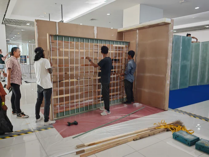 A group of workers assembling a wooden panel with a grid framework, possibly for lighting or display, as part of an exhibition booth. The setting is an indoor venue with other booth structures visible in the background. Various tools, including a drill, wooden planks, toolboxes, and a broom, are placed on the floor, which is covered with protective plastic sheets.