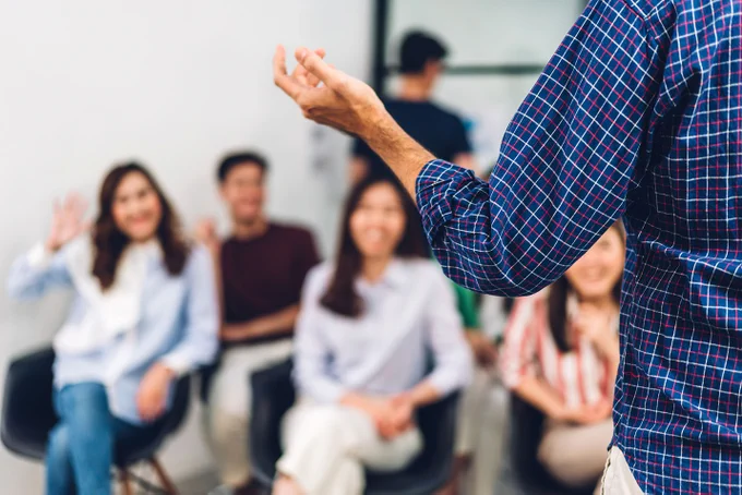 Photograph showing a lecturer in the foreground, teaching happy students who are in the background.