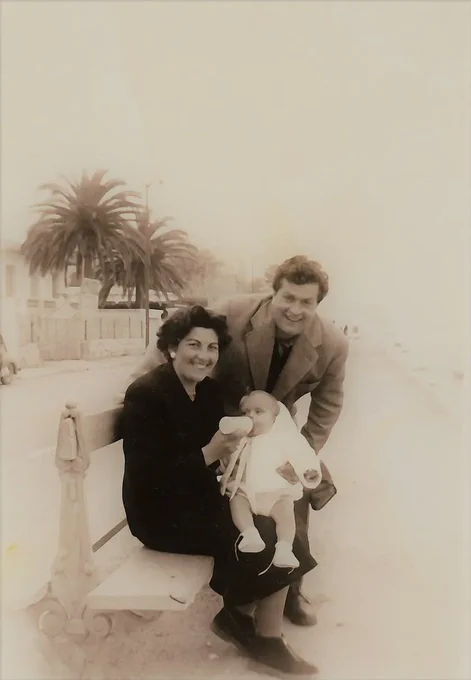 Vintage photo of two adults and a baby sitting on a bench with palm trees in the background.
