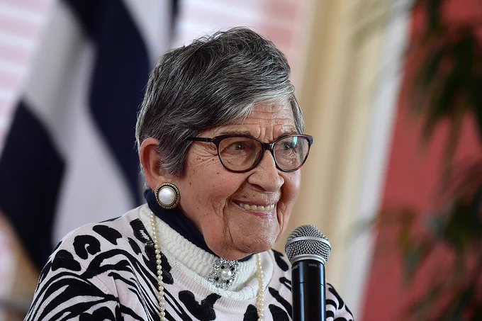 A mature woman with glasses, wearing large hoop earrings, a black and white patterned top, and a white necklace, is smiling while sitting in front of a microphone. 
