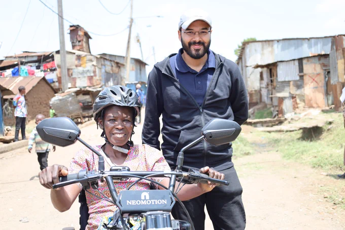 A cheerful woman wearing a helmet using a NeoMotion third-wheel mobility device, surrounded by an urban environment.