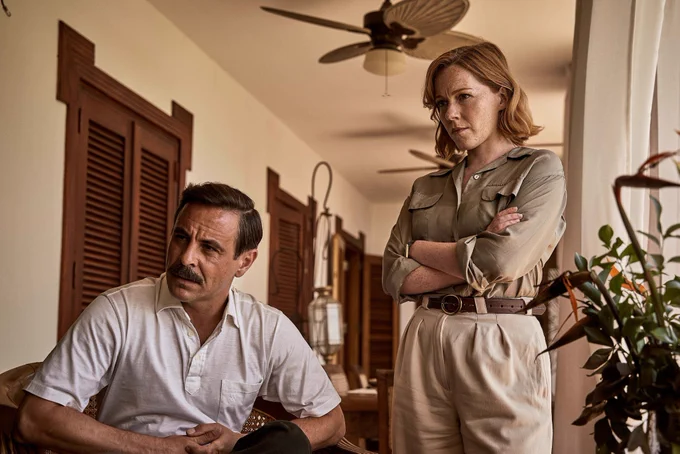 Emun Elliott and Charlotte Spencer in The Gold season 2 in a tropically styled room, both looking serious.